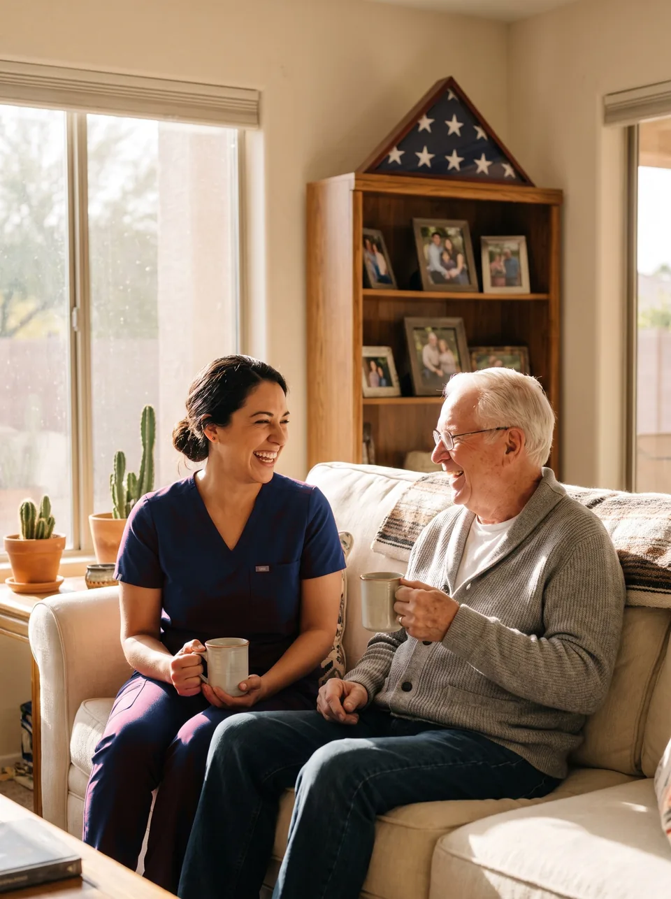 A caregiver in navy scrubs laughs with a retired man over coffee on his sofa, a folded flag in a display case on the shelf behind them